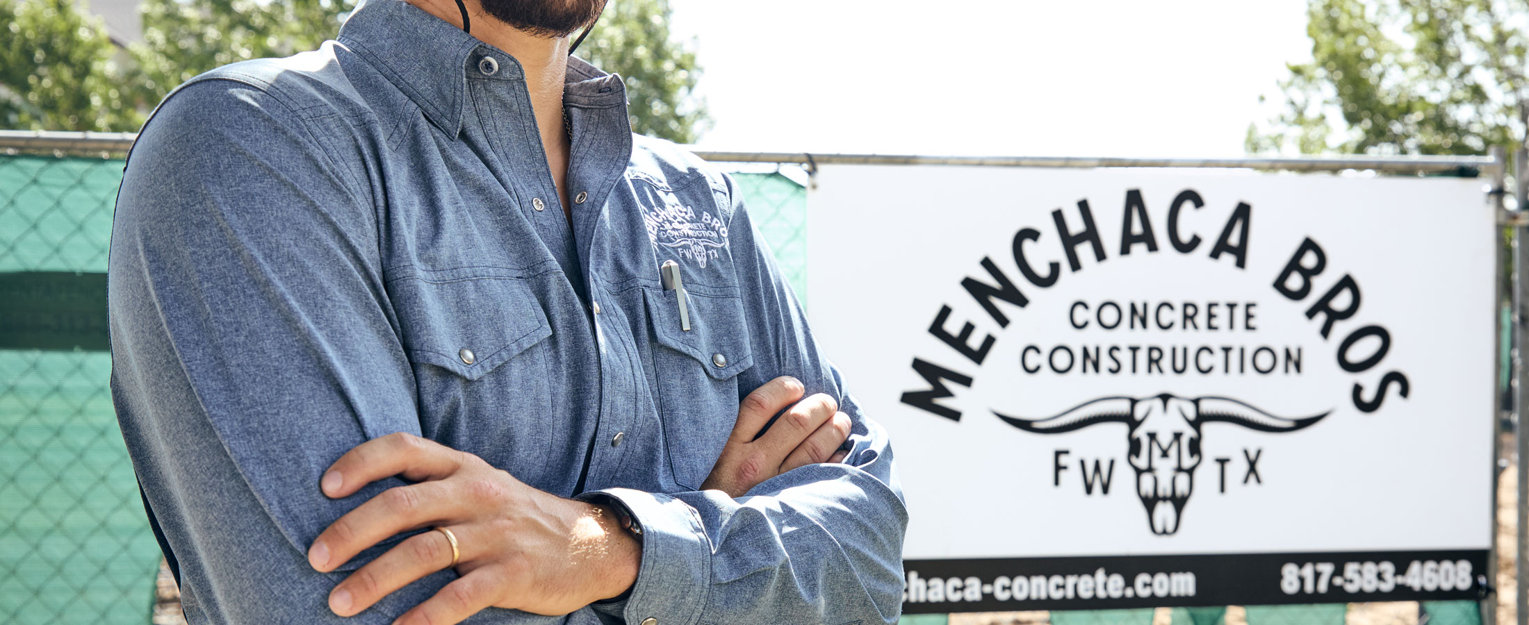 A man, Nathan Menchaca, in a blue button-up work shirt stands with arms crossed in front of a Menchaca Bros Concrete Construction sign at a job site, with fencing and trees in the background.